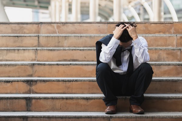 Upset stressed young Asian business man in suit with hands on head sitting on stairs. Unemployment and layoff concept.
